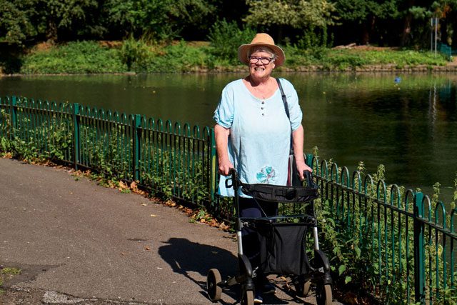 An older woman smiling while using a wheeled walker to walk around a pond
